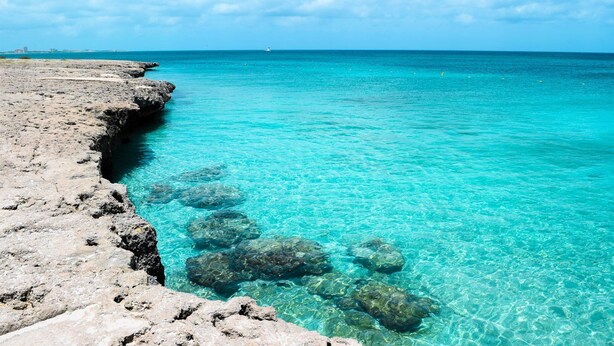 A rocky shoreline with clear turquoise water and a distant view of a city skyline. - Villa Azul - With a Jetted tub Villa 3 (savaneta)