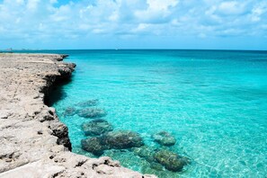 A rocky shoreline with clear turquoise water and a distant view of a city skyline. - Villa Azul - With a Jetted tub Villa 3 (savaneta)