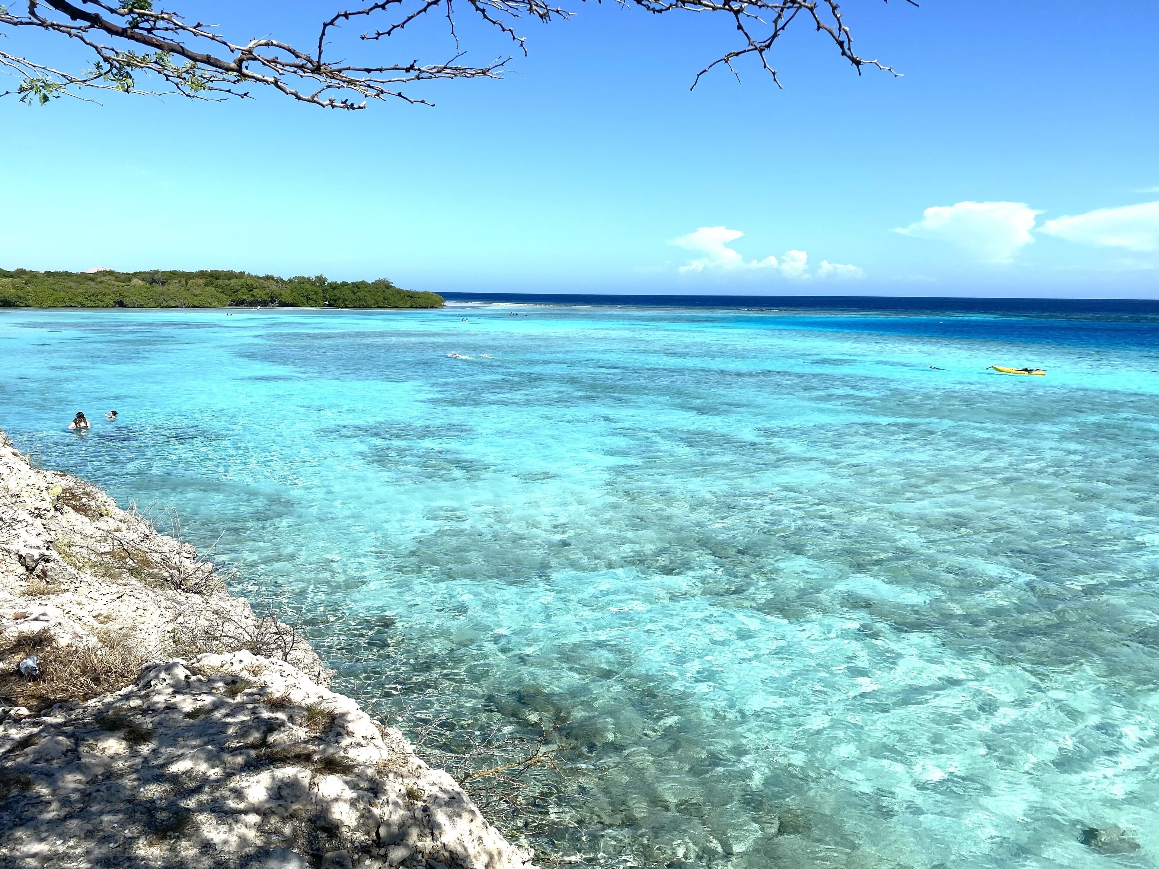 Ein freier Blick auf einen Strand mit türkisblauem Wasser, einer felsigen Küstenlinie und einer entfernten, grünen Landmasse.