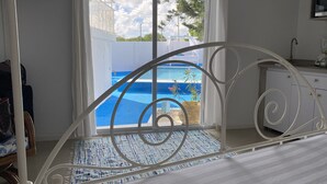 Una habitación con estructura de cama metálica, vista a una piscina y una zona de cocina con lavamanos y encimera.