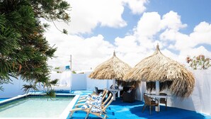 Zona de piscina con sillones de descanso y sombrillas de techo de paja, rodeada de paredes blancas y un cielo azul despejado.
