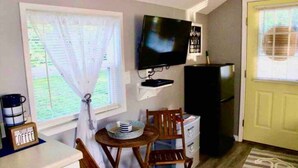 Una cocina compacta con televisor, una pequeña mesa de comedor, sillas, refrigerador y una ventana con cortinas.