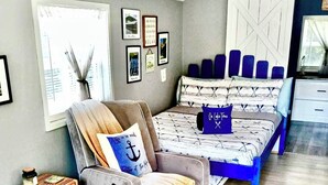 Un salón acogedor con sofá, una silla, una mesita con bandeja y una cama con cabecero azul.