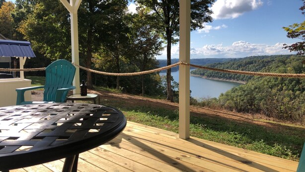 Una terraza de madera con una mesa y sillas, con vista a un lago y colinas boscosas.