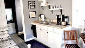 Cocina compacta con armarios blancos, un lavamanos y una cafetera. Hay una silla y una mesita con una planta.