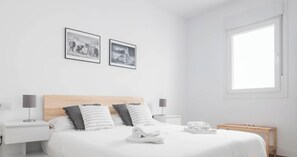 A minimalist bedroom with a wooden headboard, two bedside tables, a bed with white bedding, two pillows, a nightstand, a lamp, a window, and two framed pictures on the wall.