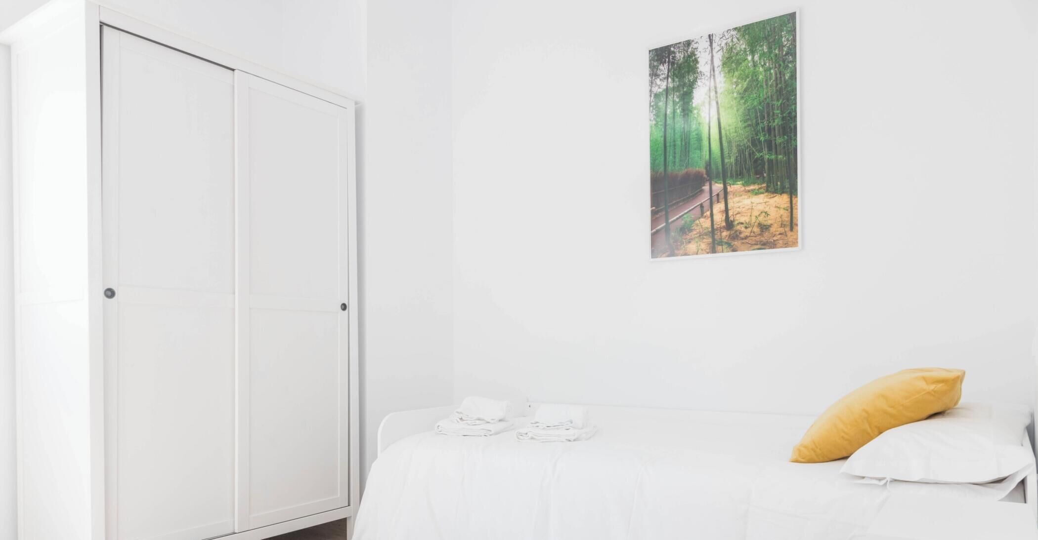 Une chambre avec une armoire blanche, un lit avec une literie blanche, un oreiller jaune et une décoration murale sur le thème de la forêt.