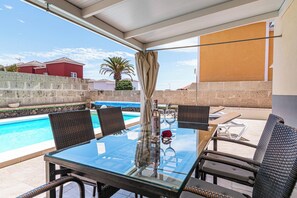 A glass-top table with wicker chairs, a bottle, and a glass set, overlooking a swimming pool.