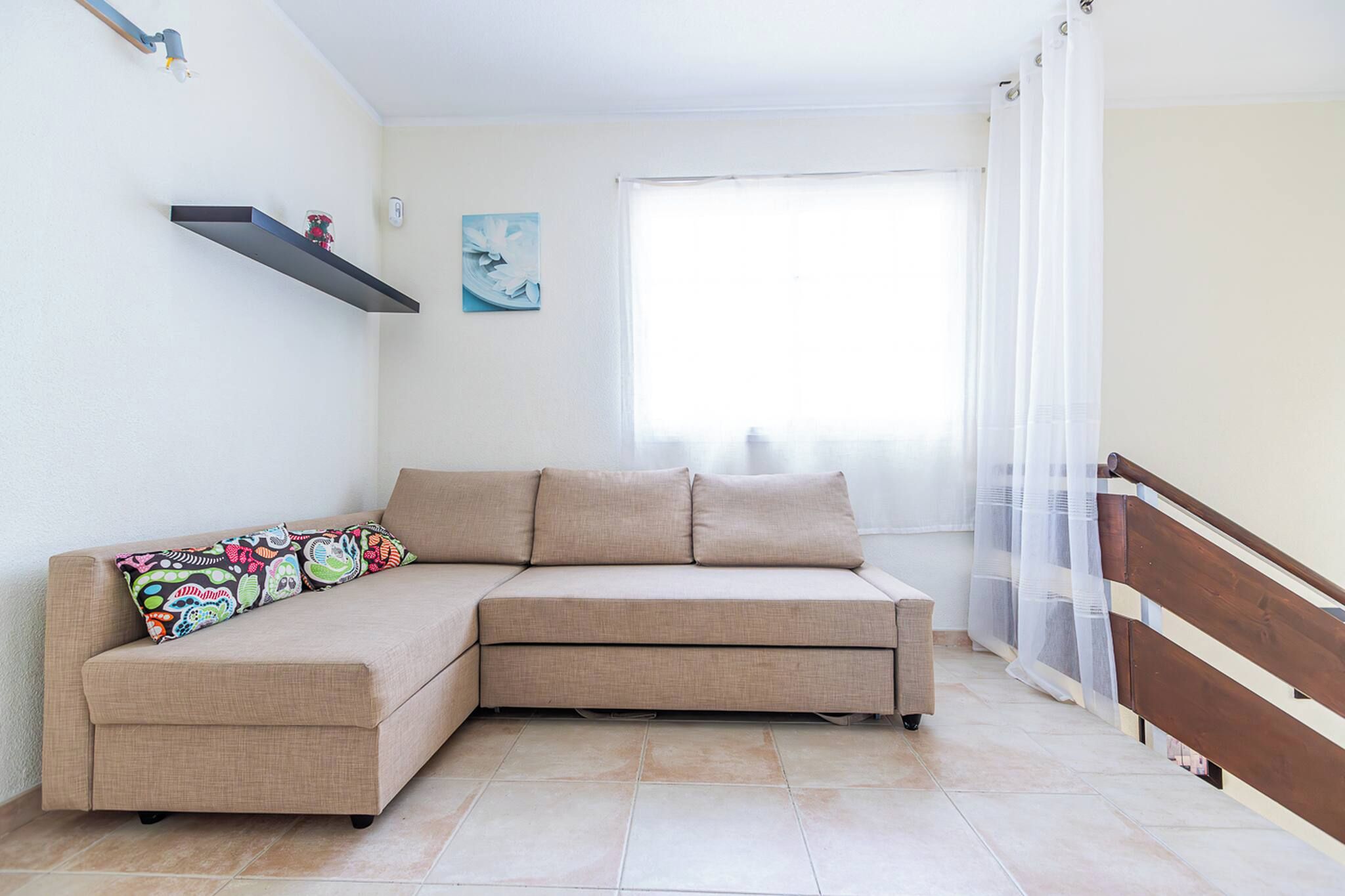 A living room with a sectional sofa, a small shelf, a painting, and a window with sheer curtains.