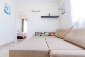 Une pièce comprenant un canapé, un lit recouvert d’une couvre-lit à motifs, une petite étagère avec des objets décoratifs et une porte donnant sur une autre pièce.