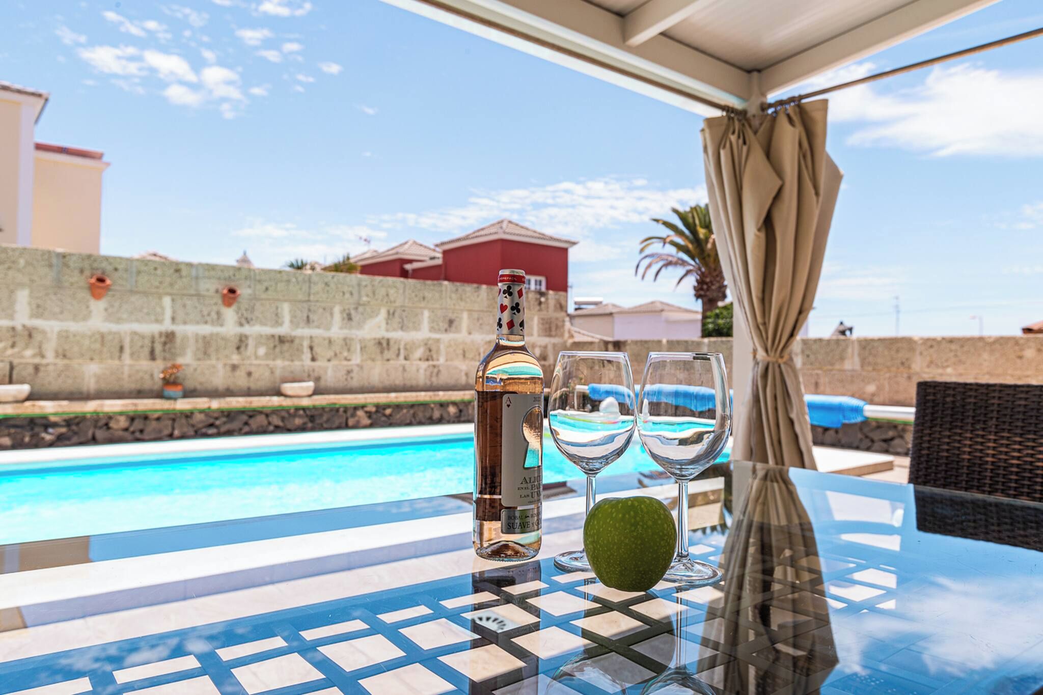 A poolside table with a bottle, two glasses, and a green fruit.
