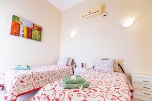 A bedroom with two beds, a wall-mounted air conditioner, a nightstand with a lamp, and a painting on the wall.