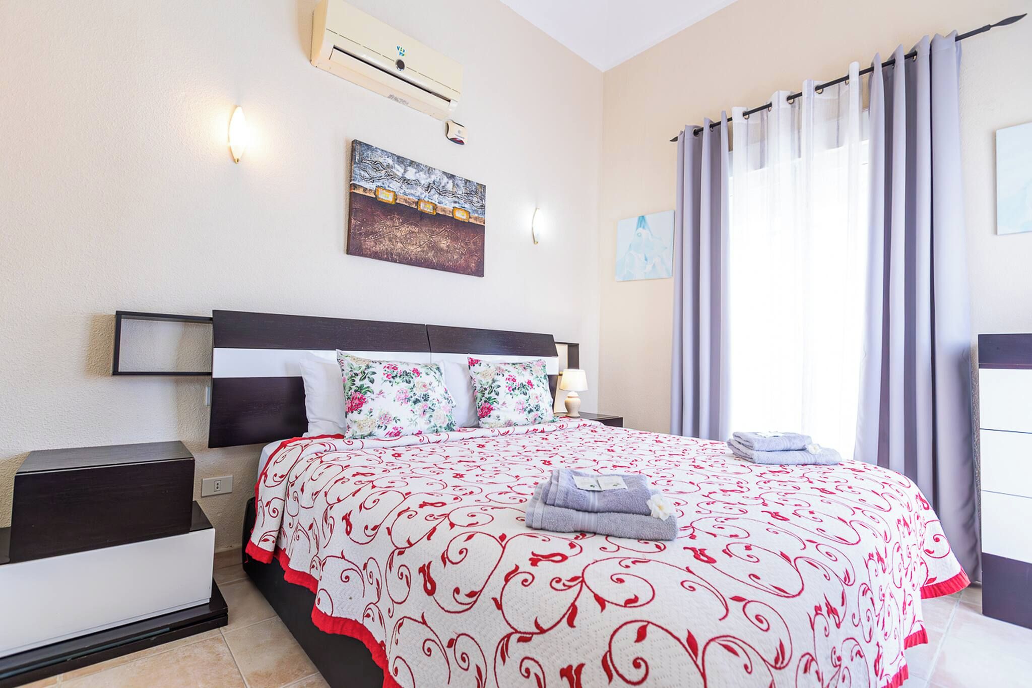 A neatly made bed with a patterned bedspread, a bedside table with a lamp, and a wall-mounted air conditioning unit.
