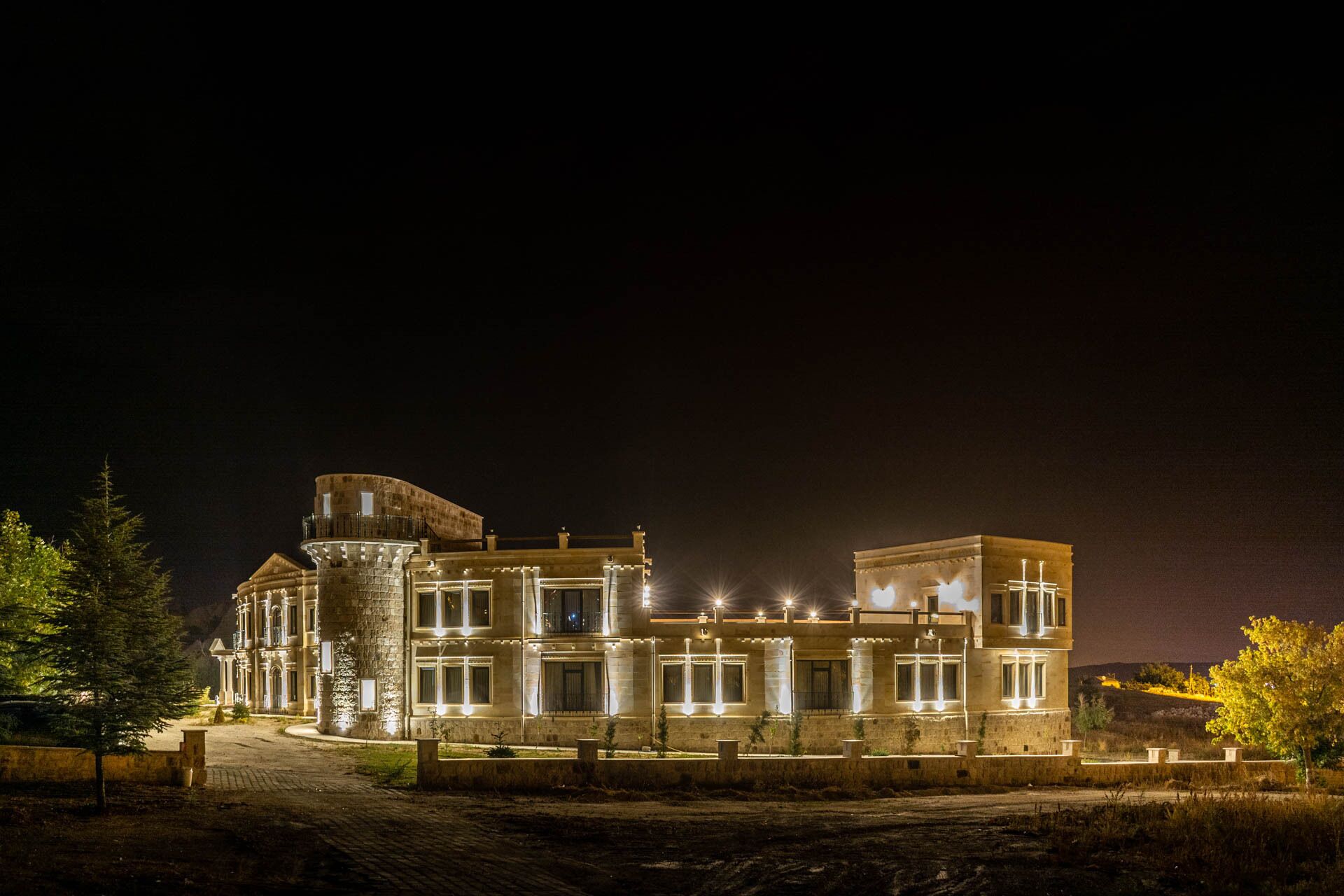 A large, illuminated building with a central tower and multiple windows, surrounded by a landscaped area.