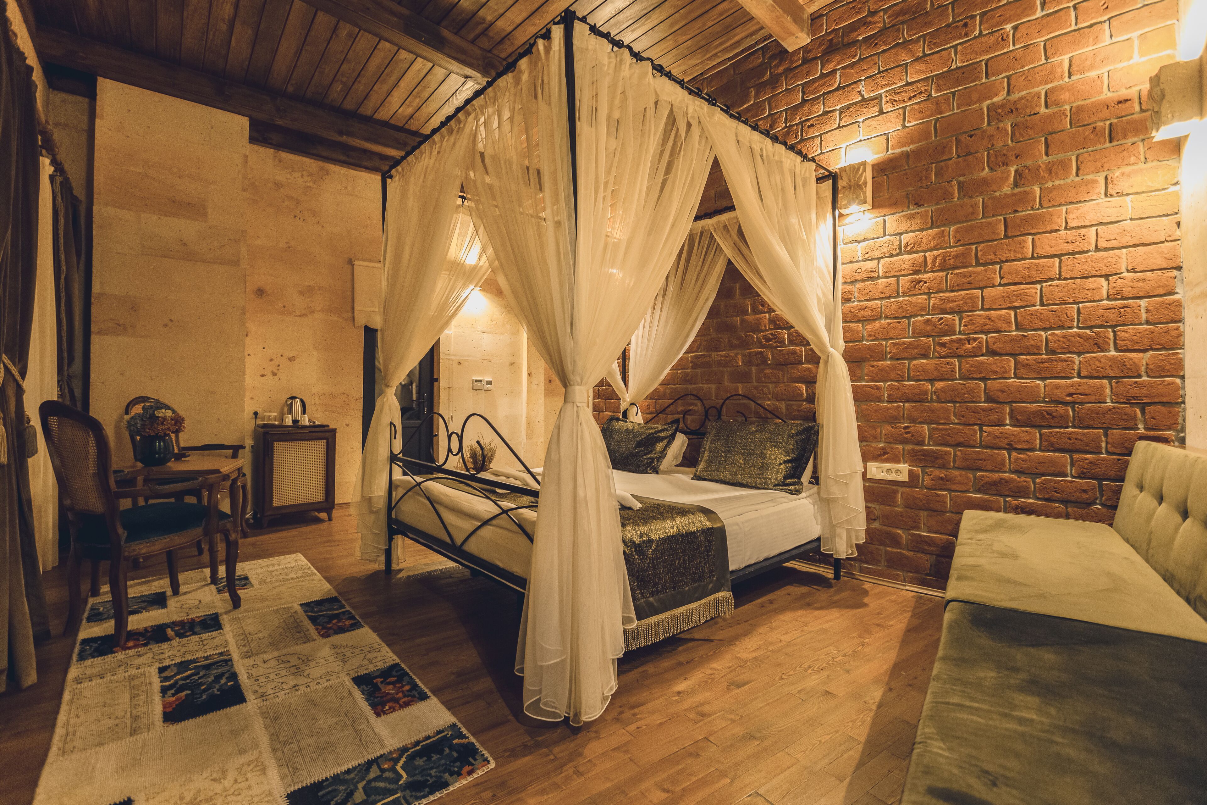 Una habitación con una cama con dosel, suelo de madera, pared de ladrillo, sofá verde y una pequeña mesa con sillas.