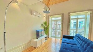 A modern living room with a blue sofa, a TV on a white stand, a floor lamp, and a large window with a view of buildings.