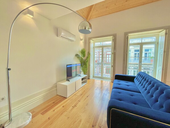 A modern living room with a blue sofa, a TV on a white stand, a floor lamp, and a large window with a view of buildings. - Porto Domus 210 Penthouse (Porto)