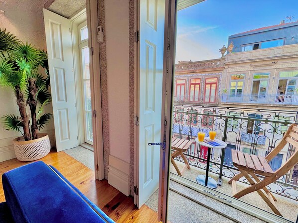 A balcony with a view of a building with balconies and windows, a table with chairs, and a potted plant.