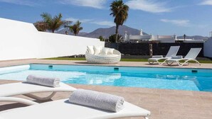Area piscina con lettini e zona relax circolare, circondata da un muro bianco e palme, con una montagna sullo sfondo.