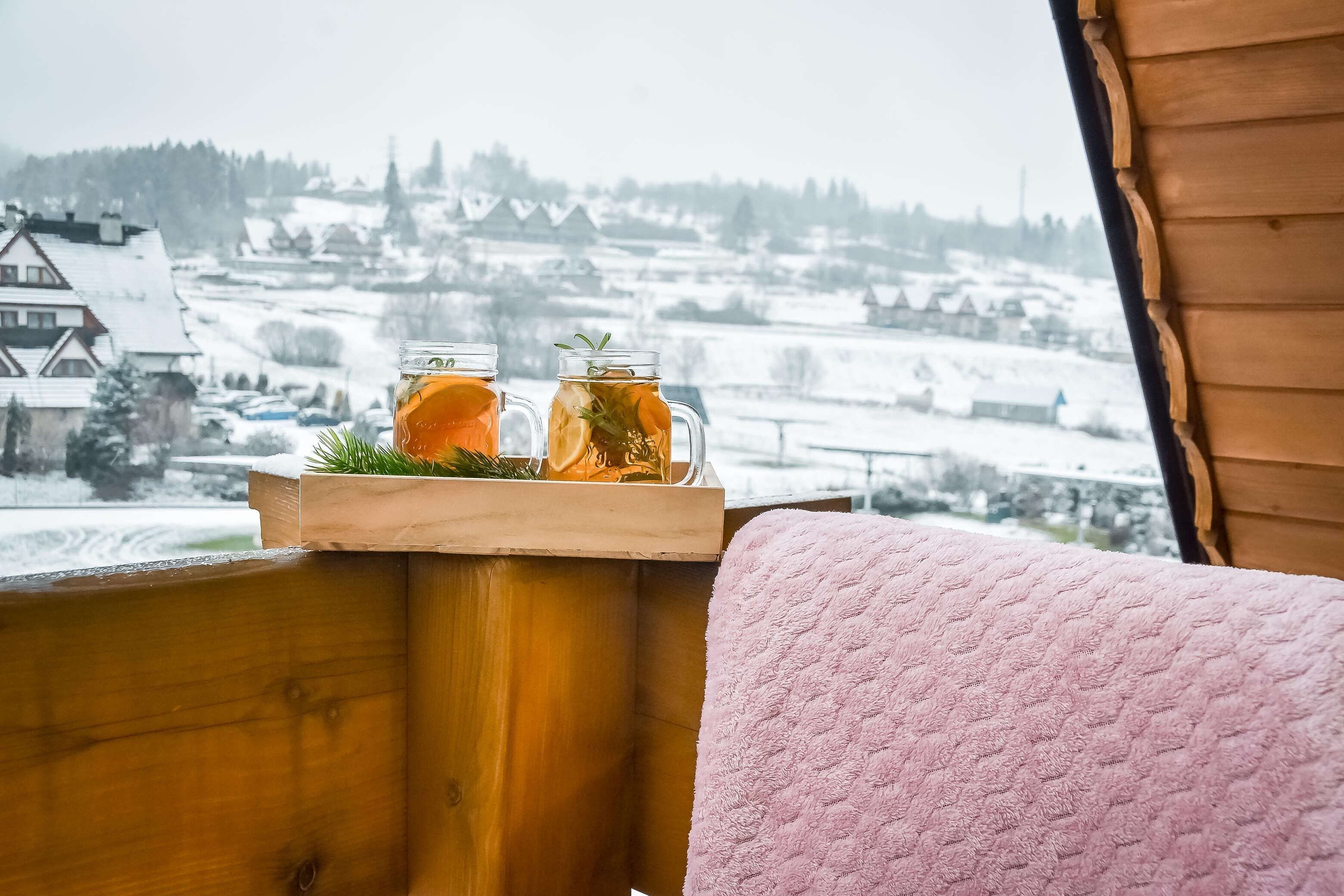 Een houten bank met een roze handdoek en twee glazen amberkleurig drankje met kruiden, met uitzicht op een besneeerd landschap.