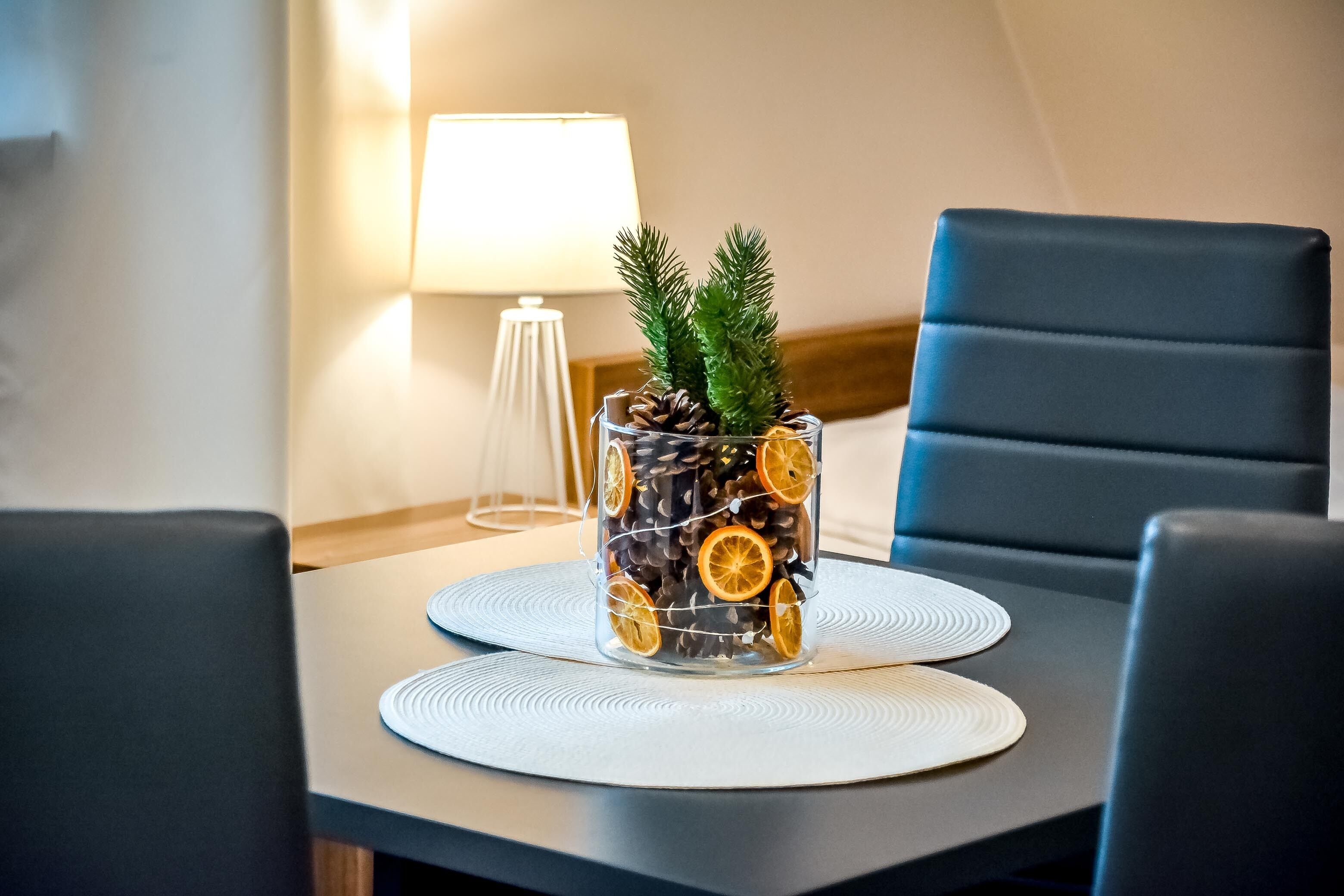 Een eettafel met een glazen vaas gevuld met dennenappels en gedroogde sinaasappelschijfjes, omringd door zwarte stoelen.