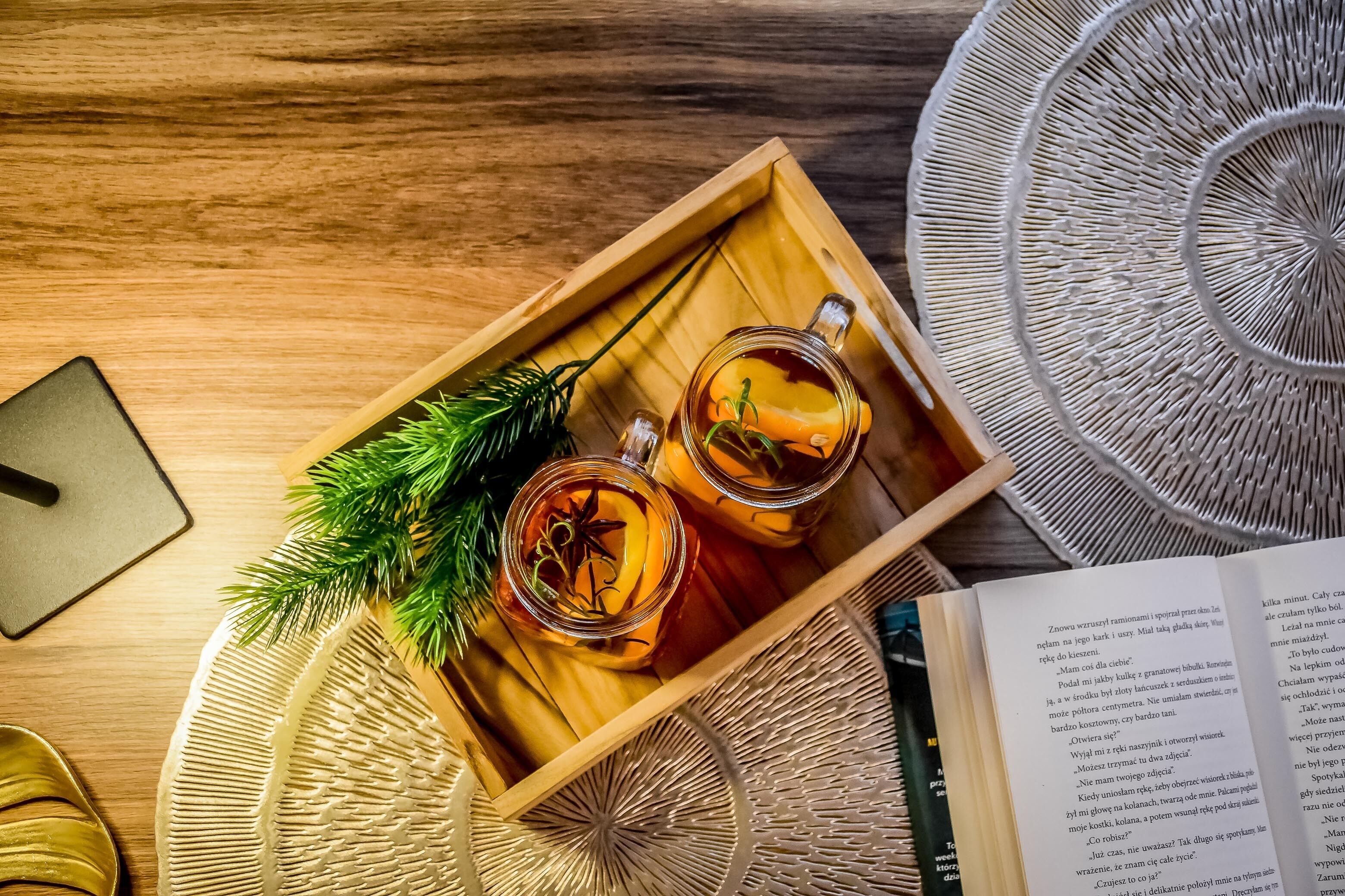 A wooden tray with two glasses of amber liquid, possibly tea, garnished with a sprig of greenery.
