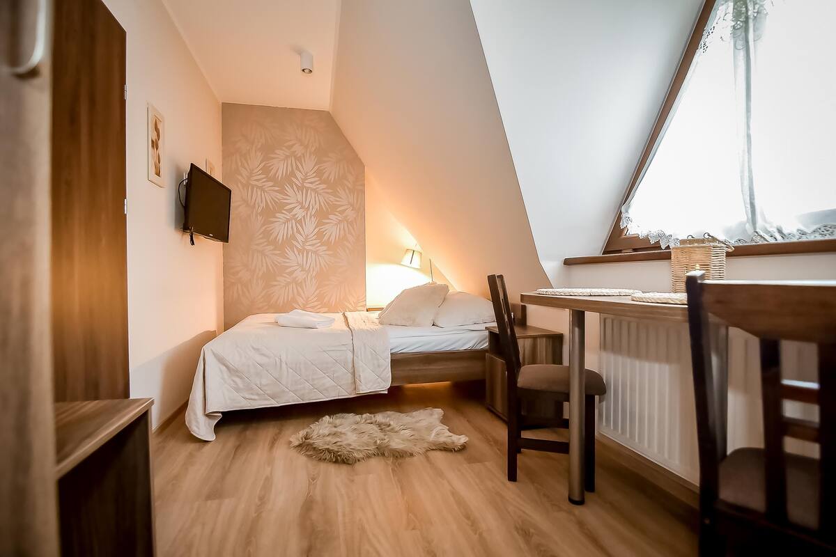 A bedroom with a bed, a desk, a chair, a television, and a window with lace curtains.