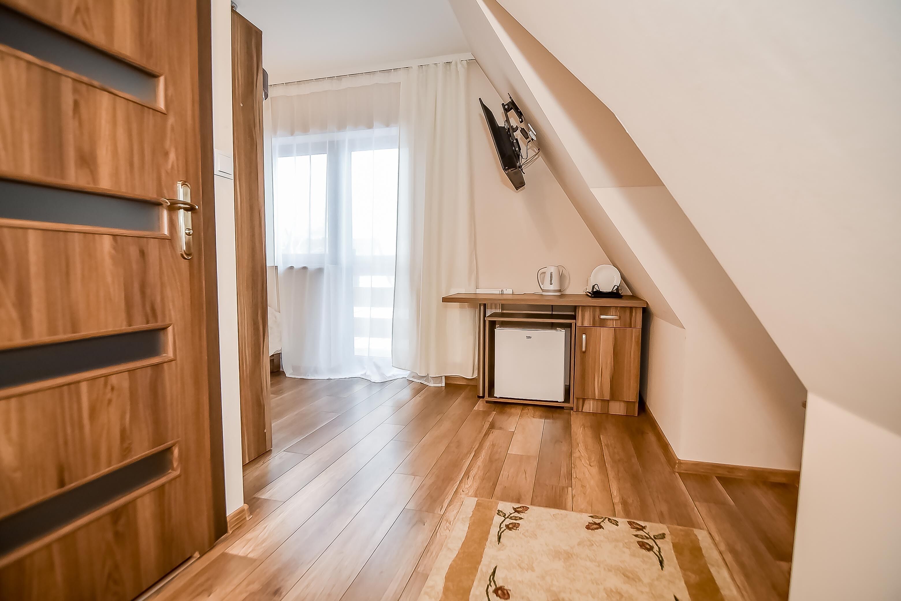 Una habitación con suelo de madera, una kitchenette pequeña con refrigerador, y una puerta que conduce a otra habitación.