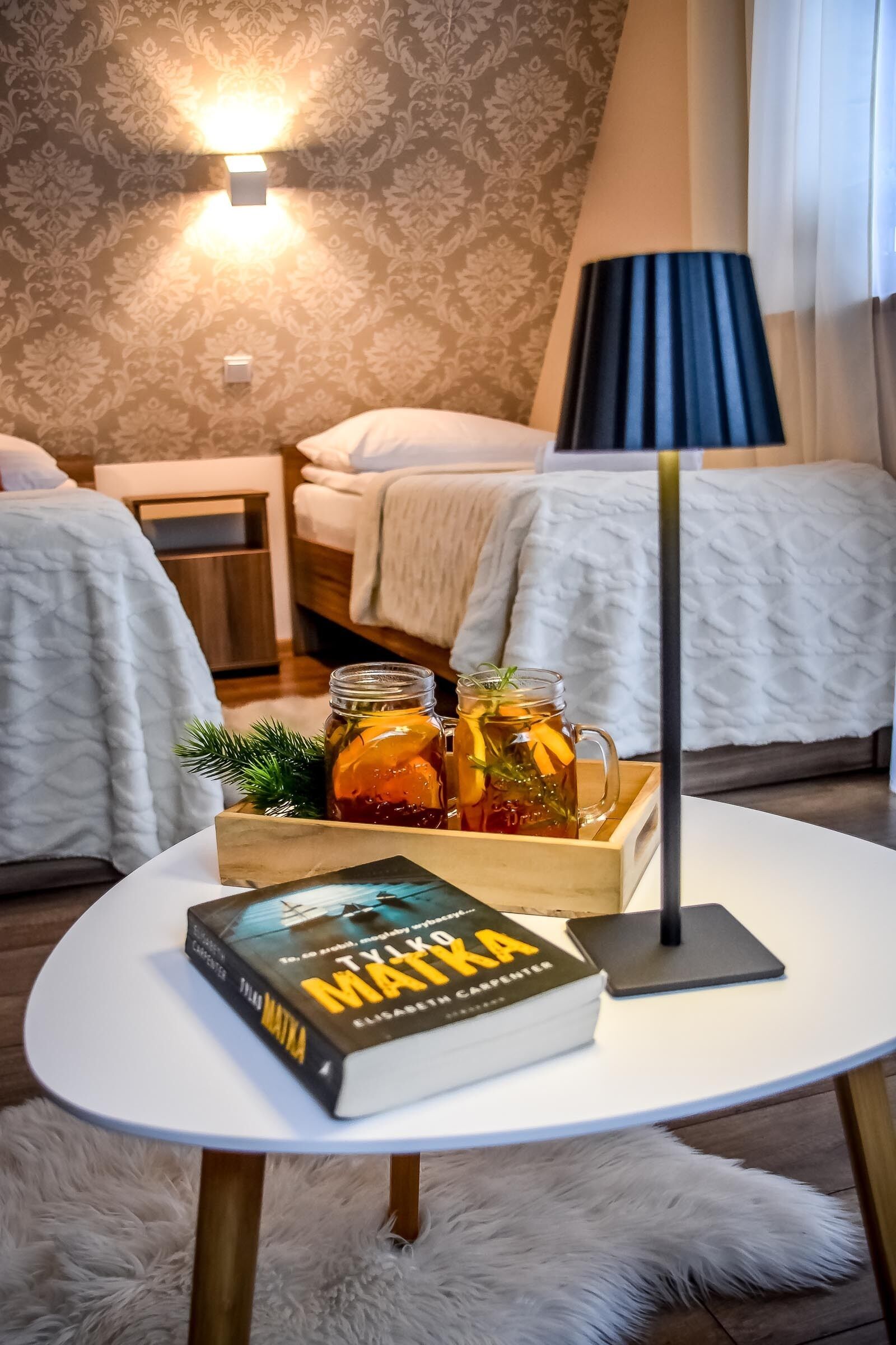 A hotel room with two beds, a small table with a book and a tray of drinks, and a lamp.