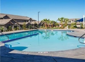 A swimming pool with clear blue water, surrounded by palm trees and a building with a sloped roof.