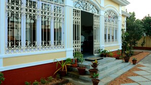 A building with ornate iron window grills, a red and yellow facade, and a variety of potted plants on the steps. - La Taara (Vanur)