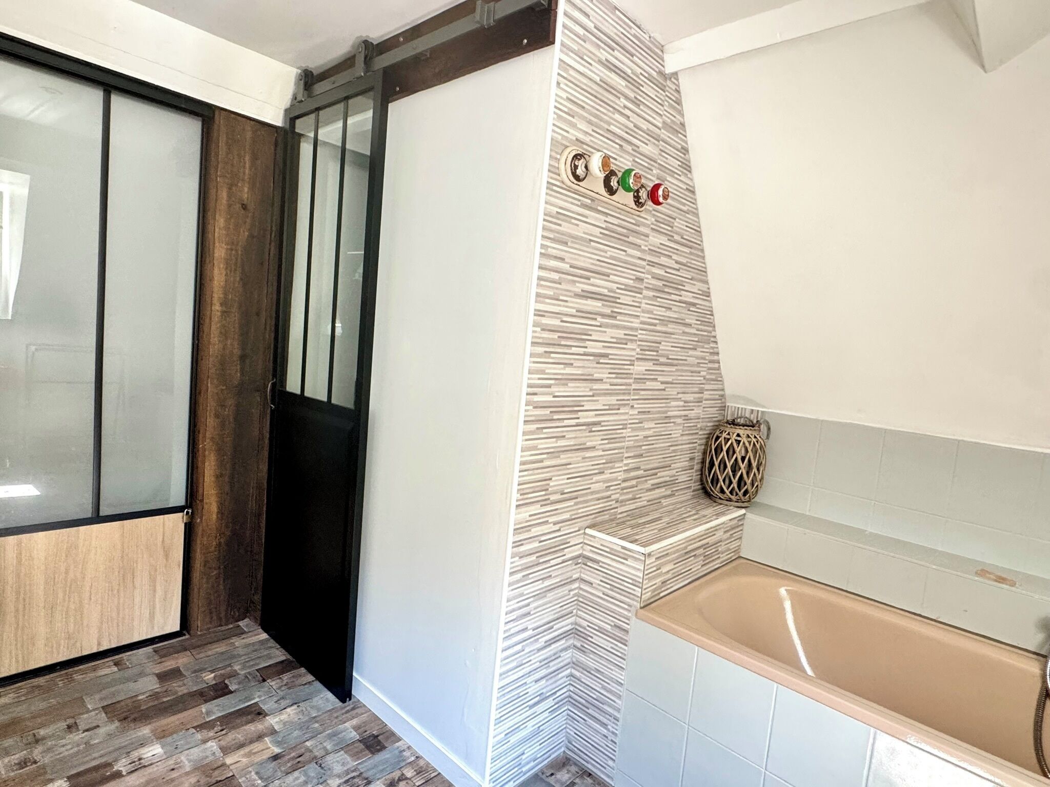 A modern bathroom with a wooden floor, a glass door, a white tiled wall, and a beige bathtub.