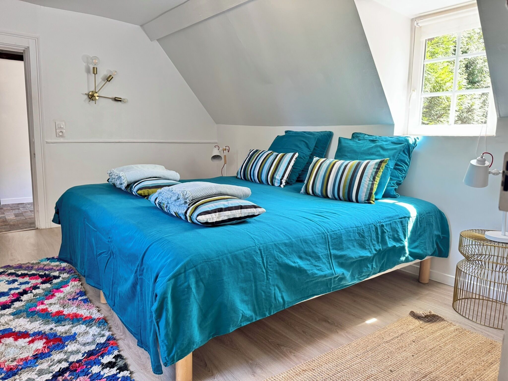Une chambre avec un grand lit, une literie de couleur turquoise, des coussins à rayures, un tapis coloré et une fenêtre donnant sur un paysage verdoyant.
