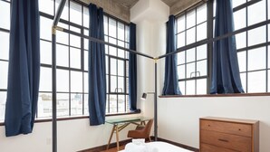 Ein Hotelzimmer mit einem Bett, einem Schreibtisch, einem Stuhl, einer Kommode und großen Fenstern mit Vorhängen.