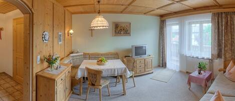 A cozy room with wooden walls and ceiling, a dining table with chairs, a sofa, a TV, and a small table with a plant.