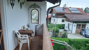 A wooden deck with a table and chairs, a white wall with decorative elements, and a garden with flowers and a small tree. - Ferienwohnung Apfelbaum 1. Etage, 42 qm, 1 Sep. Schlafzimmer, Balkon (Bad Feilnbach)