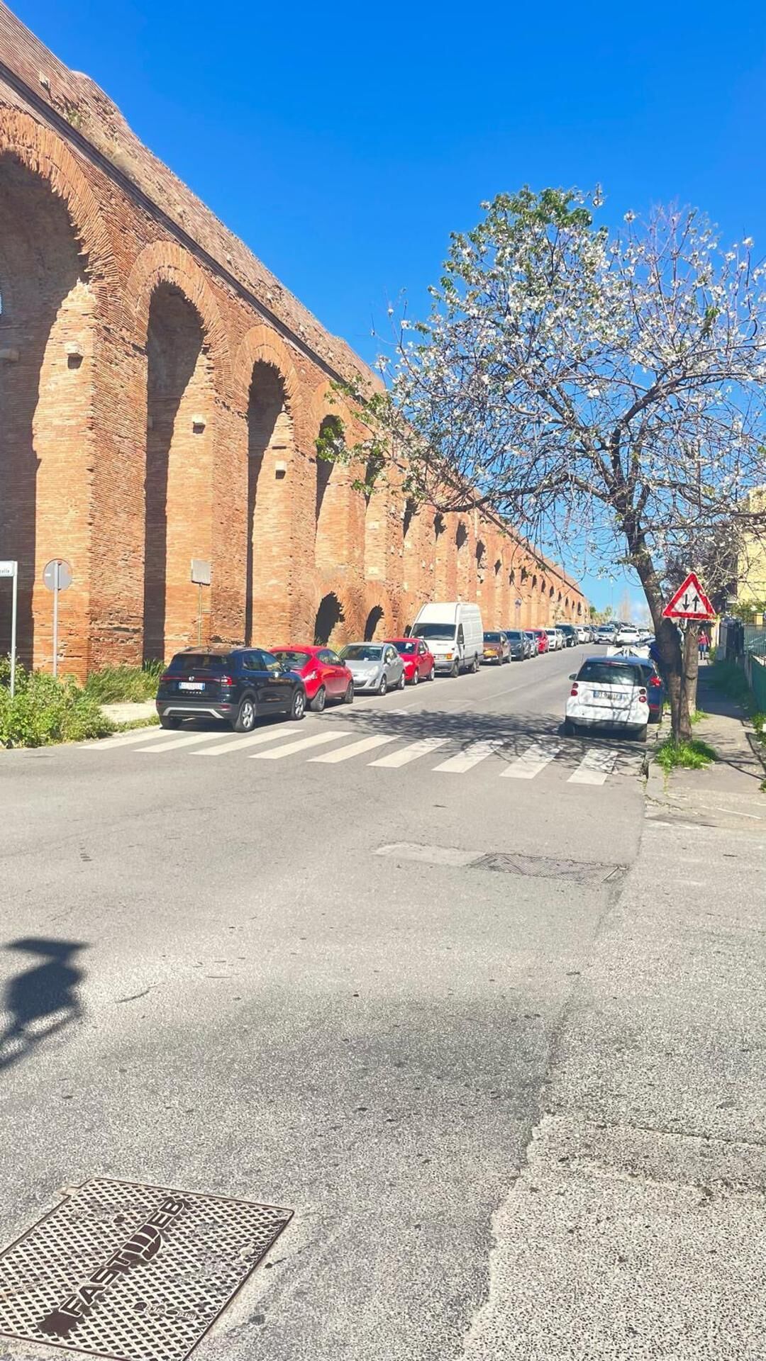 Una calle con fila de autos estacionados, un paso de peatones y un edificio grande de ladrillo con arcos.