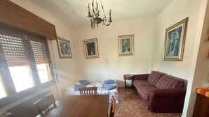 A dining area with a wooden table and chairs, a sofa, and a chandelier. There are framed pictures on the walls and a window with blinds. - Panoramic and Bright Penthouse is Waiting for you (Rome)