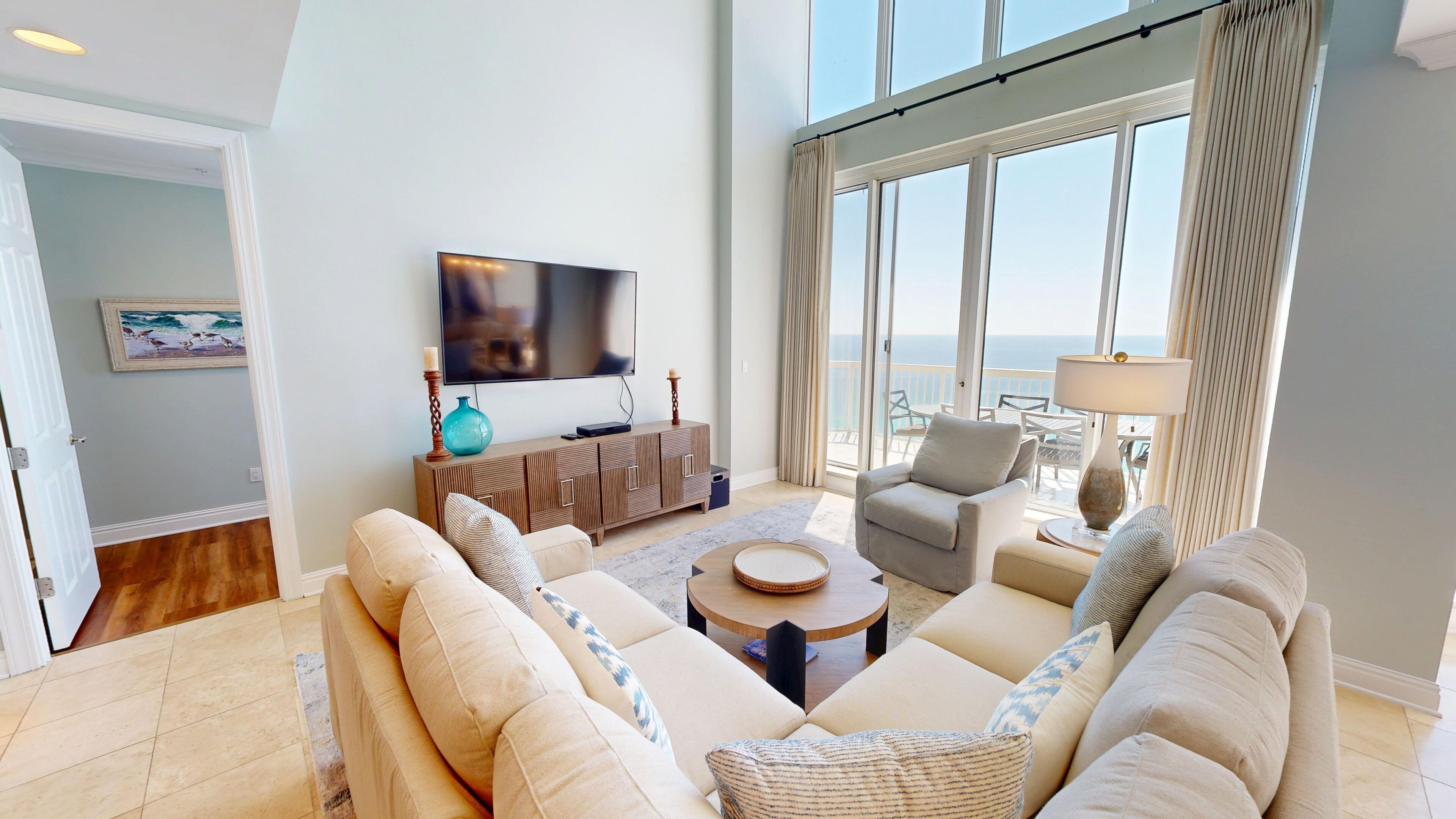 A spacious living room with a large flat-screen TV, comfortable seating, a coffee table, and a view of the ocean through large windows.