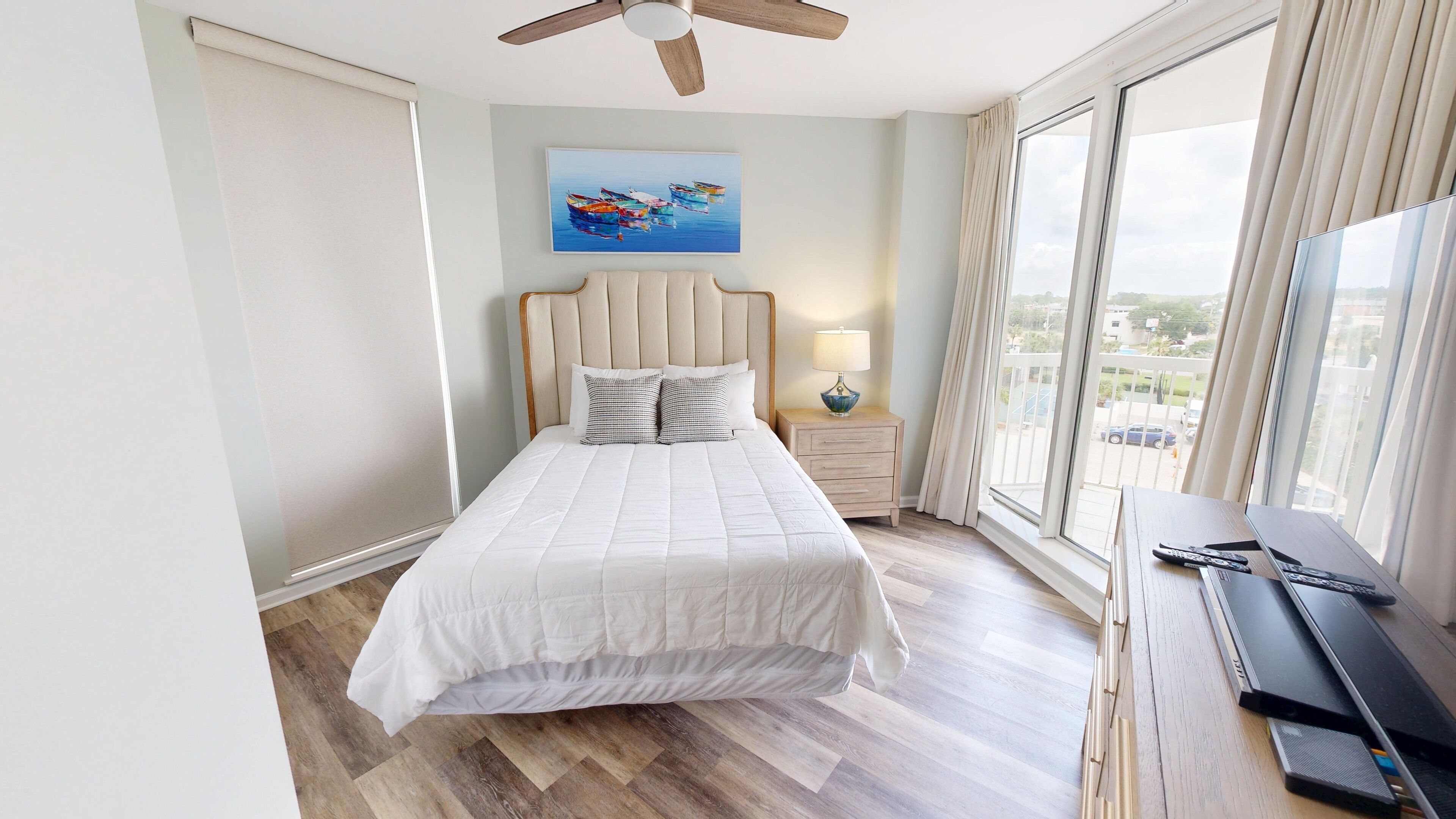 A bedroom with a bed, a nightstand, a lamp, a ceiling fan, and a large window with a view of the outside.