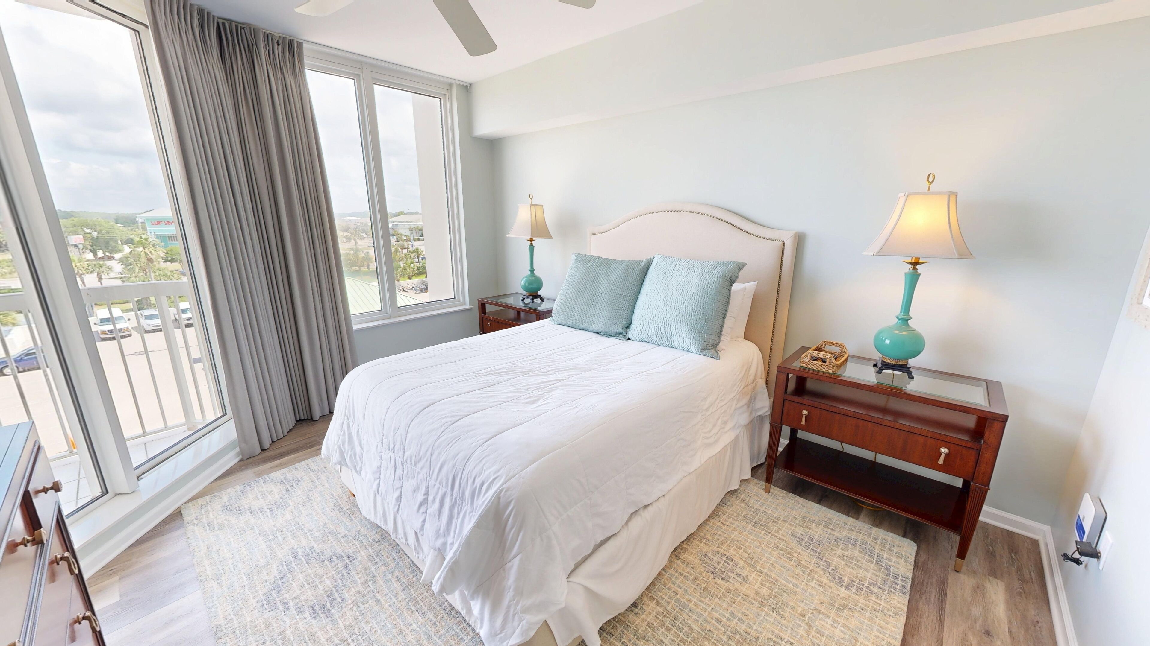 A hotel room with a large bed, two bedside tables with lamps, a view of the city through large windows, and a balcony.