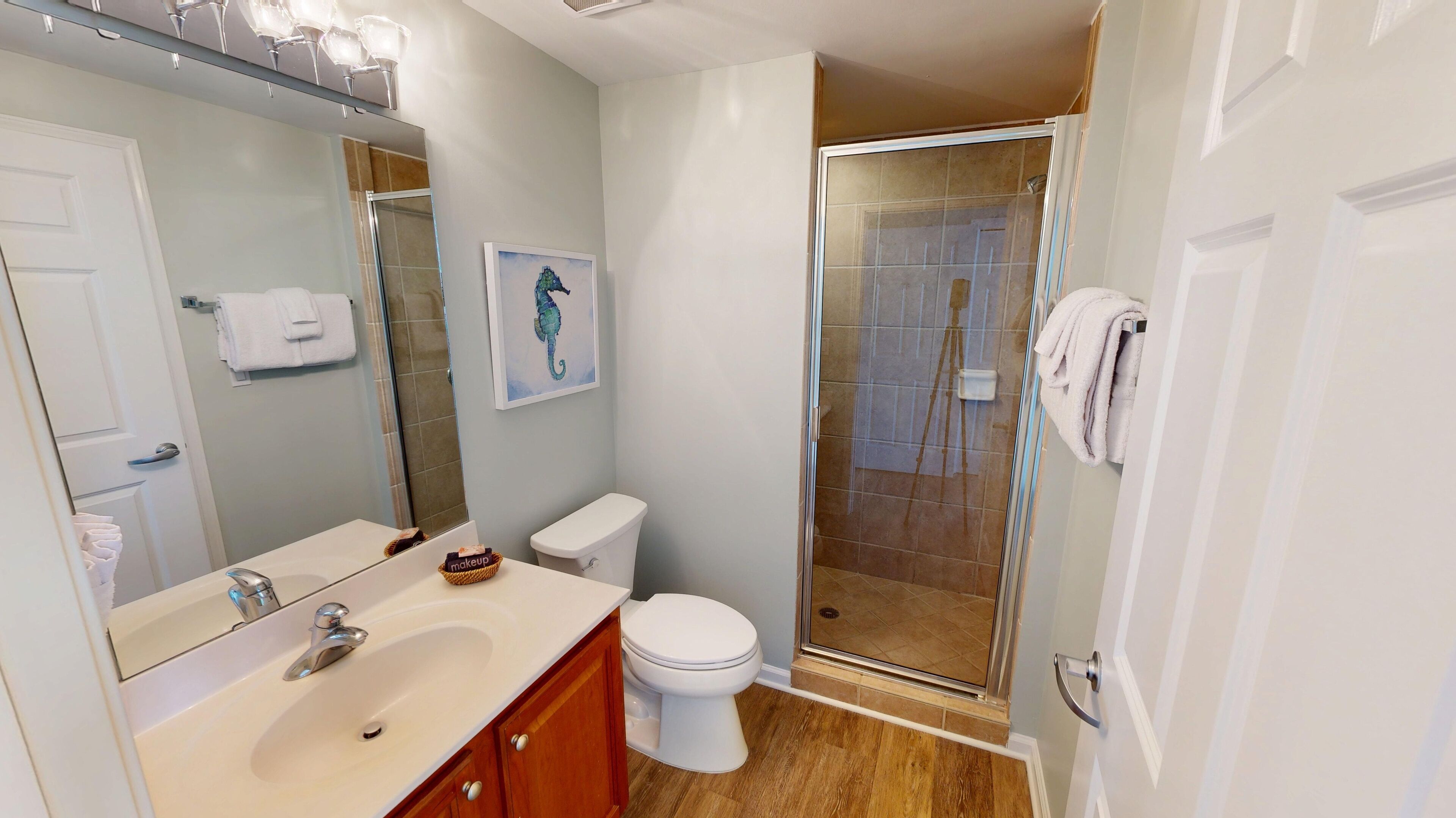 A modern bathroom with a white sink, vanity, and toilet. There is a glass shower enclosure and a seahorse artwork on the wall.