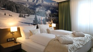 A hotel room with a large bed, two bedside tables with lamps, a wall mural of a snowy mountain landscape, and a green curtain. - Alpenlodge Val Gronda (Obersaxen)
