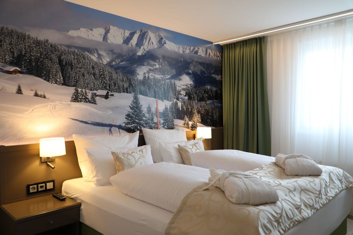 Een hotelkamer met een groot bed, twee nachtkastjes met lampen, een muurschildering van een besneeuwd berglandschap en een groen gordijn.