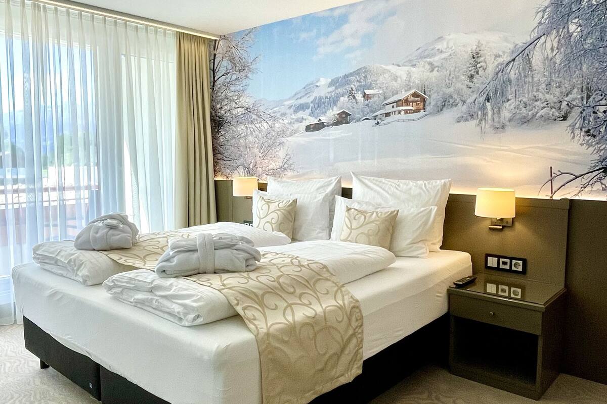 Een hotelkamer met een bed, nachtkastje, lamp en een groot raam met gordijnen.