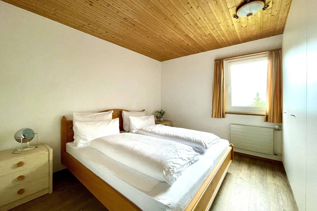 Een slaapkamer met een houten plafond, een bed met witte lakens, een nachtkastje met een lamp en een raam met gordijnen.