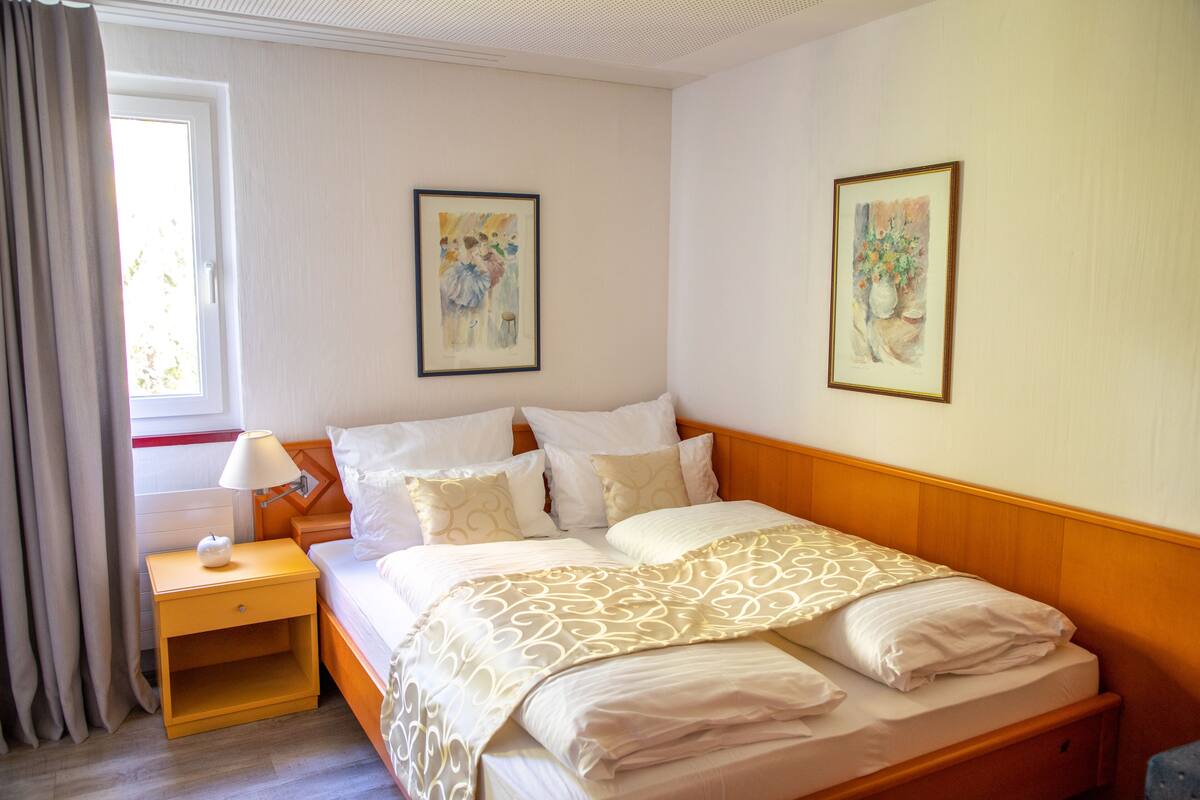Een netjes opgemaakt bed met een bedsprei met een patroon, een nachtkastje met een lamp, en twee ingelijste kunstwerken aan de muur.