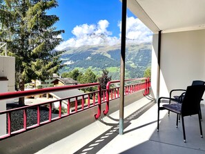 Un balcon avec une balustrade rouge, une chaise et une vue sur un paysage montagneux.