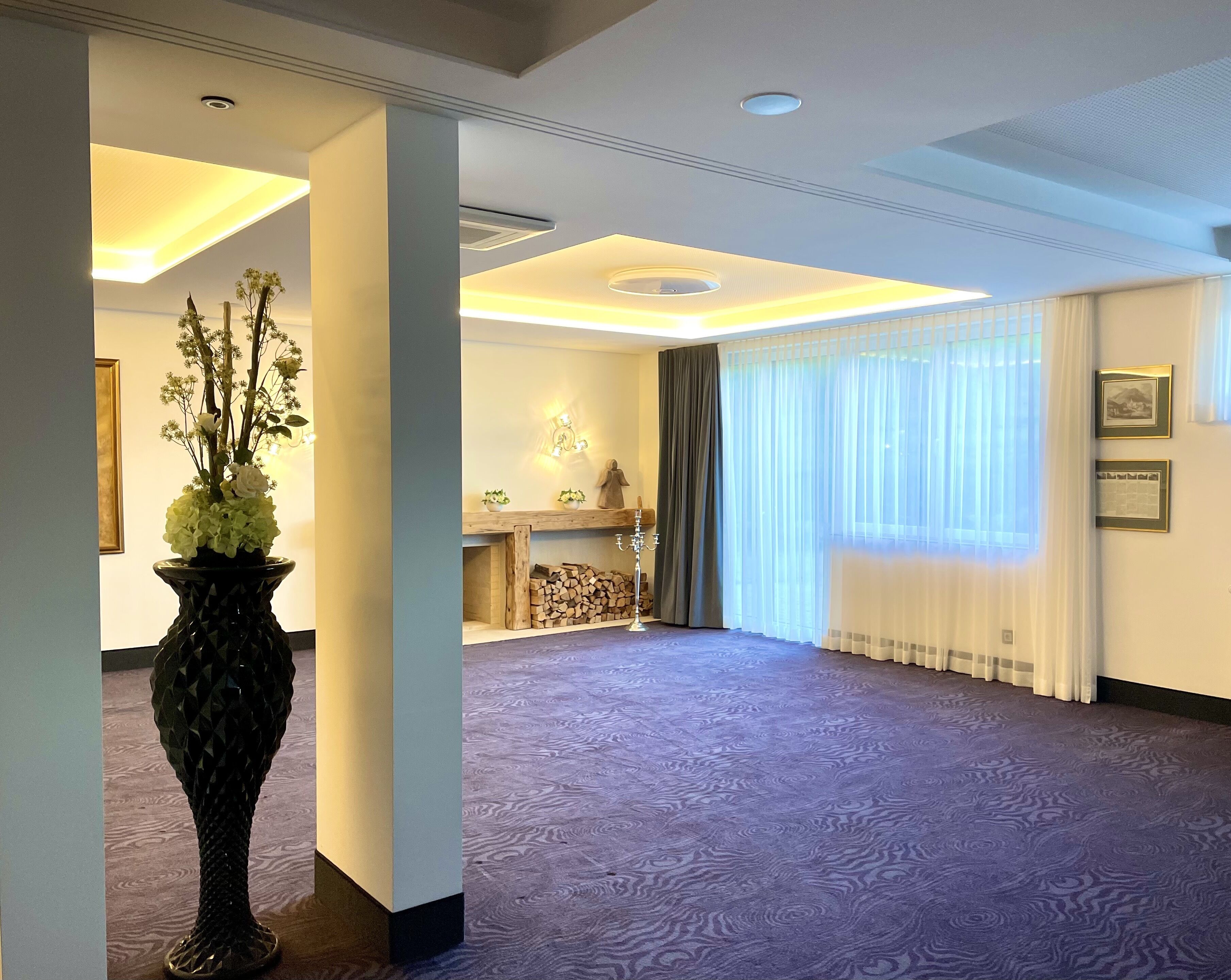 A hotel lobby with a large vase of flowers, a fireplace, and a large window with sheer curtains.