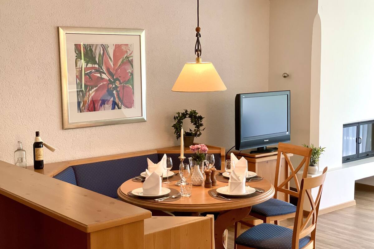 Een eethoek met een ronde tafel gedekt voor twee personen, een televisie en een lijst met een schilderij aan de muur.